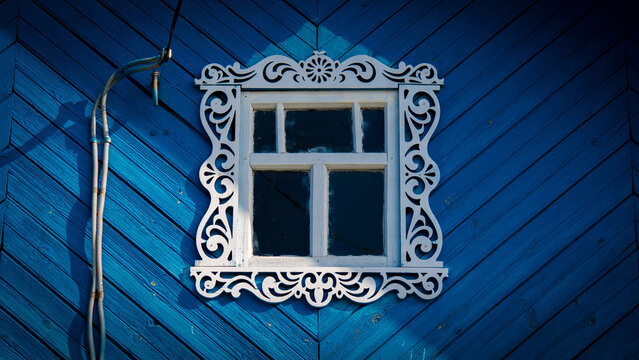 Gros plan d'une fen&ecirc;tre encadr&eacute;e avec un chambranle blanc en bois sculpt&eacute; orn&eacute; de motifs traditionnels sur la fa&ccedil;ade bleue d'une isba russe, maison construite en bois situ&eacute;e dans le nord de la Russie