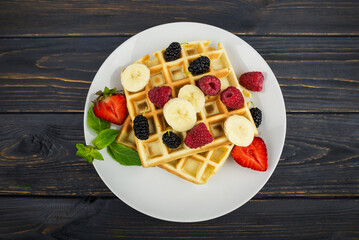 Belgian waffles with summer berries and powdered sugar in a white plate on a dark wooden background.
