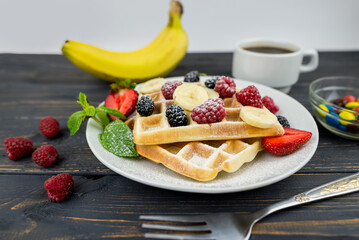 Belgian waffles with summer berries and powdered sugar in a white plate on a dark wooden background.