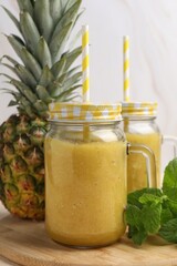 Tasty pineapple smoothie in mason jars, mint and fruit on wooden board, closeup