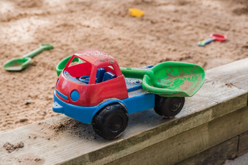 Red and blue toy truck carrying green shovel against sand pit in background, leisure and lifestyle concept illustration.