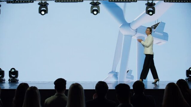 Caucasian woman female speaker presenting on the stage during ecological renewable energy conference. Future technology meetup. Talks about green energy sources, wind turbines, solar power