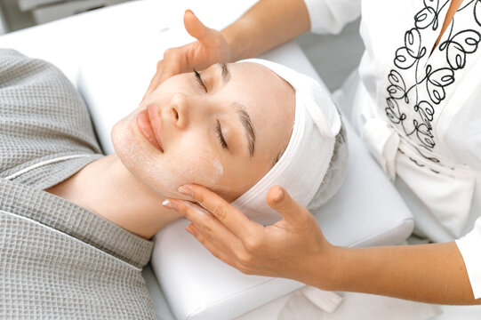 Facial skin care procedures in a beauty salon. The cosmetologist applies a special cream, scrub, peeling to the face of a pretty caucasian woman to improve the quality of facial skin. Spa day