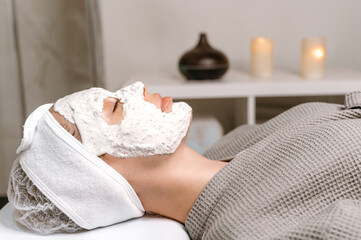 Fototapeta na wymiar Side view on a beautiful woman lying with her eyes closed in the salon with a moisturizing anti-aging mask on her face. Moisturizing nourishing facial mask, scrub, nourishing skin. Beauty procedure