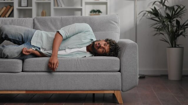 Tired Indian boy slumps on couch feeling listless. Feeling depressed, lack of motivation, sadness, boredom. Exhausted, he collapses on the couch from stress. Burn out.