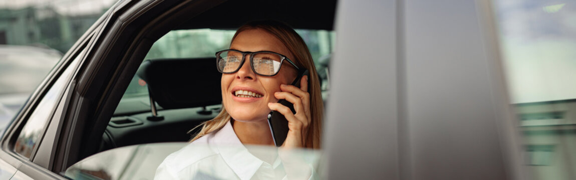 Businesswoman in eyeglasses traveling on back seat of car and talking phone while looking at window