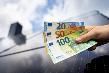 Euro banknotes in a hand with a solar panel roof in the background - obrazy, fototapety, plakaty