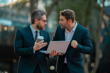 Two surprized business men team using laptop outdoor. Businessmen looking laptop with their business success in city background. Two handsome young businessmen in suits using laptop. - obrazy, fototapety, plakaty