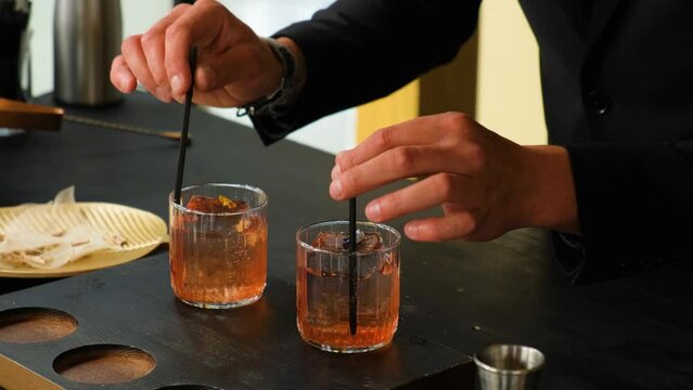 A professional bartender inserts straws into glasses with an orange cocktail and ice and serves to customers