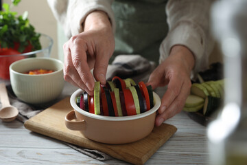 Cooking delicious ratatouille. Woman adding slice of zuccini into bowl with fresh vegetables at white wooden table, closeup