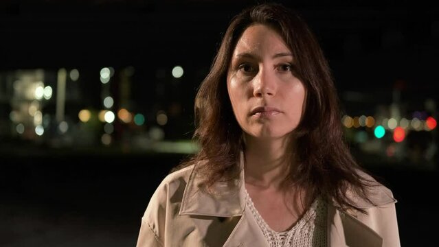 Confident young woman stands alone in the city at night and looks directly into the camera and directs a gas spray with her hand, motion blur and replay