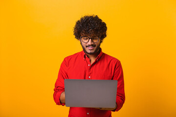 Portrait of cheerful young brunette man with beard typing email on laptop and smiling, reading...