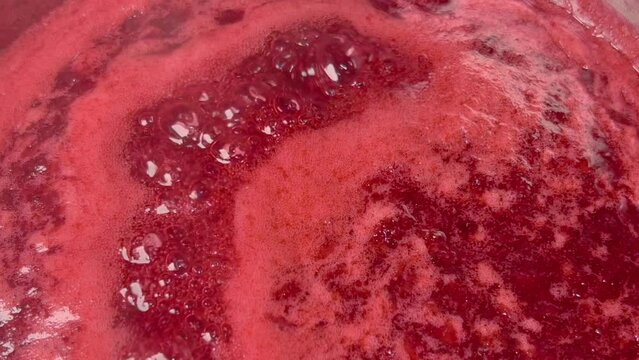 Boiling red jam is cooked in a saucepan. Air bubbles and steam slowly come to the surface.