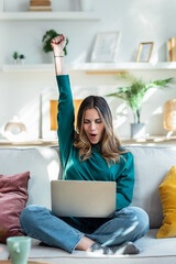 Pretty young woman celebrating something while using her laptop on the sofa at home.