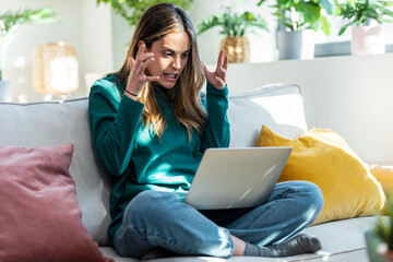 Angry frowning young woman working online on laptop sitting on couch at home