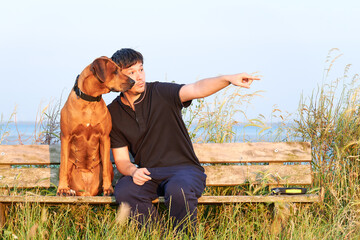 Young man sitting outdoors with his Rhodesian ridgeback dog on wooden bench and pointing finger at something ahead 