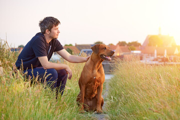 Young man sitting outdoors with his Rhodesian ridgeback dog at summer evening