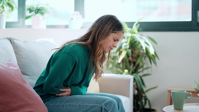 Video of unhealthy young woman with stomachache leaning on the couch in the living room at home.