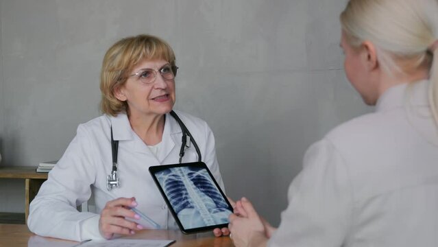 An elderly blonde doctor in a white coat, sitting at her desk, shows the patient the results of a chest X-ray on an electronic tablet and points with a pen to the details of the image. High quality 4k