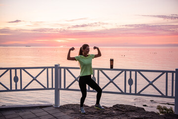 Fototapeta premium Young woman is fooling around in warm-up in morning at dawn of sun by sea. Sportswoman in sportswear