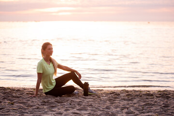 Fototapeta na wymiar Young woman in sportswear is sitting on the seashore at sunrise. Girl is resting after workout