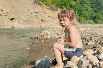 Fototapeta na wymiar The boy came ashore from the cold water of the mountain river and basks in the sun