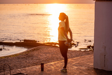 Young woman in sportswear is warming up early in morning on seashore at sunrise. Fitness