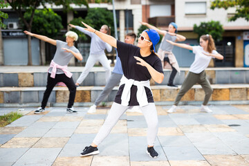 Positive girl dancing modern choreography with group of teenagers on city street on summer day.
