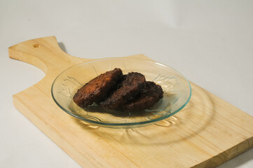 Three pieces of tempe bacem are served on a small plate on a wooden board isolated on a white background.