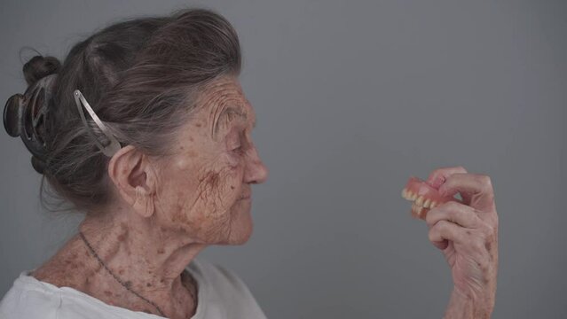 Pensioner keeps dental implants in hand and watches it against a gray backdrop in studio. Elderly woman holding her dentures in her arm. Senior citizen female looks at her dental. Tooth implants. 