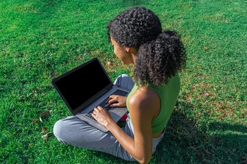 Chica joven sentada sobre el césped con un ordenador portátil sobre las piernas, trabajando, estudiando y conectada con los amigos. - obrazy, fototapety, plakaty