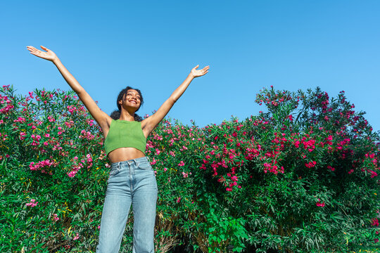 Chica joven contenta levantando los brazos mandando un gran abrazo a la humanidad, con un fondo vegetal de parque con flores y cielo azul, un d&iacute;a soleado de primavera.
