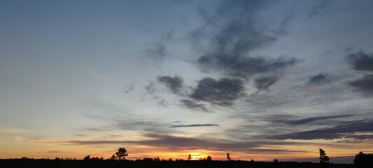 Spring sunset over the field. The sun has gone below the horizon, its last rays are visible. There are high gray clouds in the blue sky. Below them is a dark field and the outlines of growing trees.