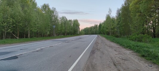 Country asphalt road. On a spring evening, a gray strip of asphalt highway goes into the distance. Along the edges of the road grows green grass and tall trees with green foliage.