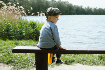 Toddler boy sitting on bench near pond. Little boy enjoys summer walk in countryside. Children outdoor activities.  - obrazy, fototapety, plakaty