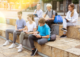 Focused teen students making notes in workbooks during lesson outside school in warm autumn day.