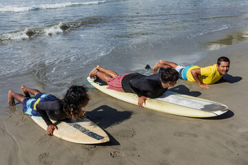 latin surf instructor and two men beginner surfers try to stand up on surfboard on lesson at beach...