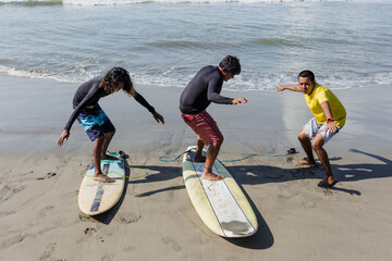 latin surf instructor and two men beginner surfers try to stand up on surfboard on lesson at beach...