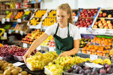 Obraz premium Hardworking fifteen-year-old girl who works part-time in a store as a trainee saleswoman puts grapes on the counter, laid ..out in boxes for sale