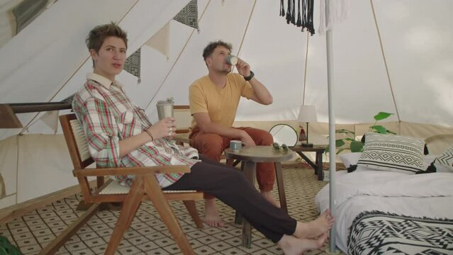 Full shot of young man and woman sitting in glamping tent, having tea and speaking while spending vacation together