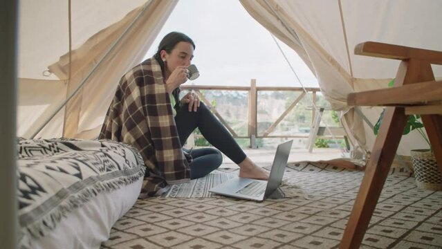 View from inside of glamping tent of girl sitting on floor, using laptop and drinking tea while spending vacation in nature