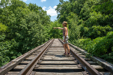 Naklejka na ściany i meble Caucasian woman on railroad tracks in the country woods on a sunny day pointing into the distance.