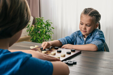 Cute boy playing checkers with little girl at table in room, closeup