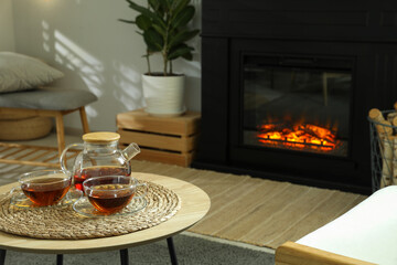 Teapot and cups of drink on coffee table near stylish fireplace in cosy living room. Interior design