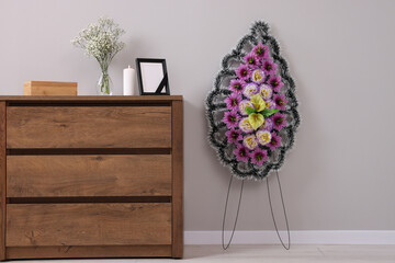 Wreath of plastic flowers and frame with black ribbon, burning candle on commode in room. Funeral...