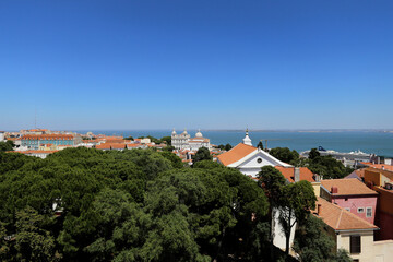 Uma deslumbrante vista aérea de Lisboa em um dia ensolarado, onde se mesclam áreas verdes, belas construções e o azul do mar ao fundo. - obrazy, fototapety, plakaty