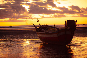A fishing boat on the sandy beach in Jericoacoara, Ceará, during sunset. The sky is adorned with vibrant shades of orange, reflecting on the sea and wet sand.