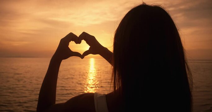 Silhouette of lovely woman Making a Heart Sign Gesture and Holding Hands Up looking at ocean sunset. Love sign concept. Rear view female make Love heart shape from hands at sunset. Summer vacation