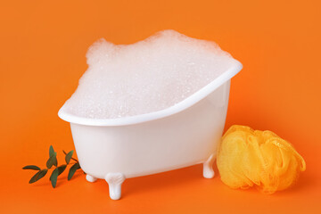 Small bathtub with foam, sponge and eucalyptus branch on color background