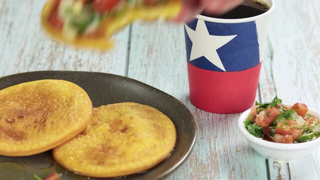 Sopaipillas with bite, hand grabbing, of squash or pumpkin and placing pebre, in round plate with polypaper cup with Chilean flag, on wooden table, Horizontal video. concept of Chilean national holida